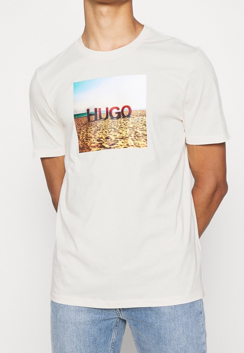 Homme portant un t-shirt blanc avec une scène de plage rectangulaire et le texte "HUGO" imprimé sur la poitrine, associé à un jean bleu clair.