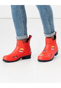 Bottes en caoutchouc rouges jusqu'à la cheville avec des motifs de canard et des parapluies, affichant une semelle extérieure bleu marine et des panneaux élastiques sur les côtés pour un enfilage facile.