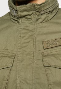 Olivgrön jacka med hög krage, dragkedja framtill och två bröstfickor. Har sydda detaljer och en tygpatch med text.