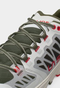 Κοντινό πλάνο παπουτσιού πεζοπορίας σε ελιά πράσινο και λευκό με γκρι κορδόνια, κόκκινες λεπτομέρειες ραφής και επάνω μέρος από διαπερατό πλέγμα.