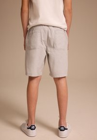 Persona che indossa pantaloncini bianchi e sneakers Adidas Stan Smith bianche, in piedi con la schiena rivolta verso la fotocamera su uno sfondo beige.