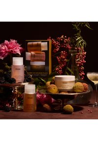 Zestaw do pielęgnacji włosów OUAI z szamponem, żelem do mycia ciała i peelingiem do skóry głowy, prezentowany wraz z jeżynami, owocami liczi, różowymi kwiatami i kieliszkiem szampana.