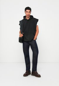 Junger Mann in ärmellosem schwarzen Hoodie, dunklen Jeans, braunen Schuhen, der eine schwarze Umhängetasche hält und vor einem weißen Hintergrund steht.