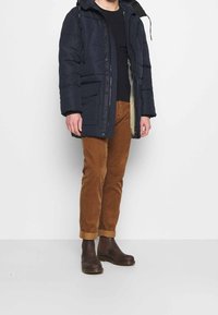 Puffjacke in Marineblau mit Reißverschluss und Druckknopfverschluss, ausgestattet mit Kapuze, Seitentaschen und beigem Futter. Getragen mit braunen Cordhosen und braunen Stiefeln.