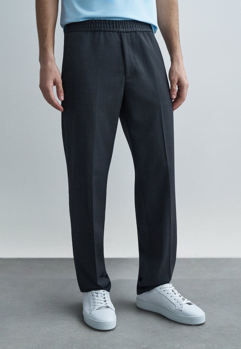 Tumedat halli värvi püksid elastse vööosaga, sirge säärega disain ja peene tekstuuriga, sobitatud valgete tossudega.