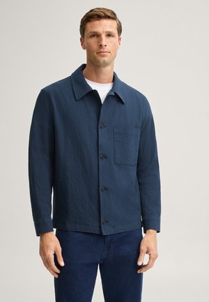 Mann trägt eine strukturierte dunkelblaue Jacke mit Knöpfen über einem weißen Hemd und dunkelblauen Hosen vor einem schlichten hellen Hintergrund.
