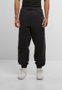 Sorte sweatpants med elastisk talje og manchetter, der har en strukturfinish og en diskret sort grafisk detalje på venstre ben, parret med hvide sneakers.