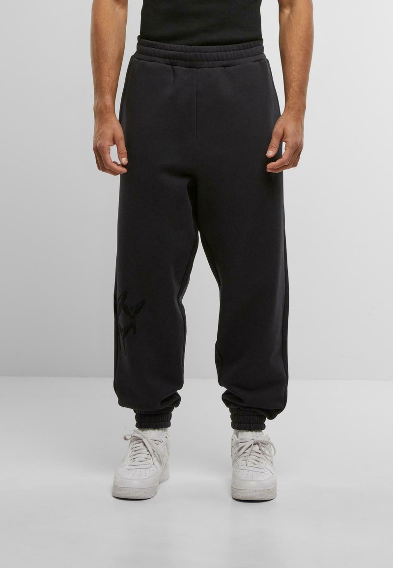 Sorte sweatpants med elastisk talje og manchetter, der har en strukturfinish og en diskret sort grafisk detalje på venstre ben, parret med hvide sneakers.