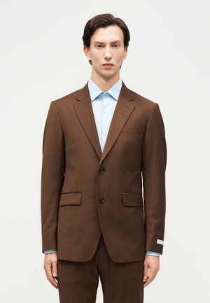 Άνδρας που φορά καφέ σακάκι δύο κουμπιών και παντελόνι που ταιριάζουν, με ανοιχτό γαλάζιο πουκάμισο με κουμπιά, στέκεται μπροστά σε μια απλή φόντο.