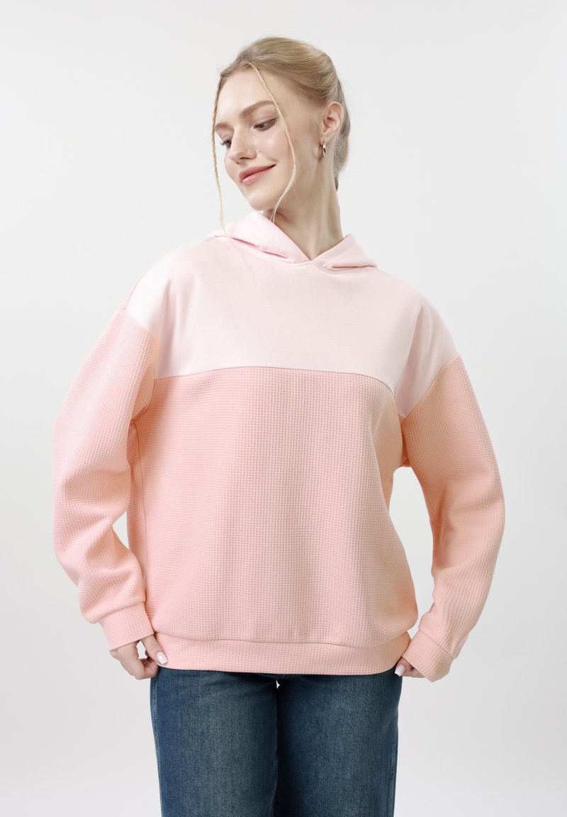 Jeune femme portant un sweat à capuche ample bicolore rose avec un tissu texturé, associé à un jean bleu, debout devant un fond blanc uni.