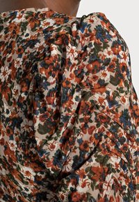 Blomstret bluse med bølgede mønstre i mørk orange, blå og grøn på en lys baggrund. Har blødt, tekstureret stof med draperingsdetaljer.