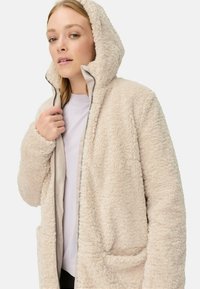Cremfarbener Fleece-Jacke mit Kapuze, mit einem Reißverschluss, weichem Material und Fronttaschen. Ungefüttertes Innenfutter mit sichtbaren Nähte. 