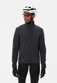 Giacca softshell nera con colletto alto, tasca sul petto con zip e dettagli riflettenti. Indossata con guanti neri e pantaloni da ciclismo aderenti. Presenta un logo carattere.