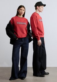 Κόκκινο φούτερ με λογότυπο "tommy", συνδυασμένο με σκούρο τζιν παντελόνι με φαρδιά μπατζάκια. Ένα μοντέλο φοράει μαύρο μπουφάν τύπου bomber, ενώ το άλλο φοράει μαύρο καπέλο.