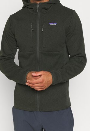Man in een donkergroene Patagonia-hoodie met rits en borstzakken, handen gevouwen voor zich, marineblauwe broek zichtbaar aan de onderkant.