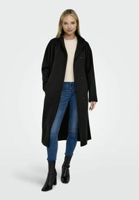 Manteau long en laine noire mélangée avec un large col, fermeture à boutons et fentes latérales, associé à un haut en tricot crème, un jean bleu et des bottes noires à talons.