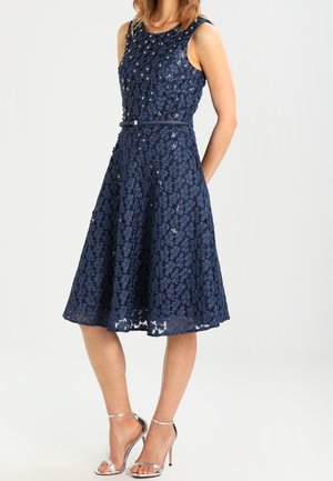 Donkerblauwe kanten jurk met een getailleerde bovenkant, uitlopende rok en bloemenpatroon. Heeft een smalle riem in de taille en een mouwloos ontwerp.