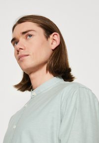 Camicia azzurra chiara con una texture morbida e colletto alla cinese, caratterizzata da una chiusura a un bottone vicino al collo.