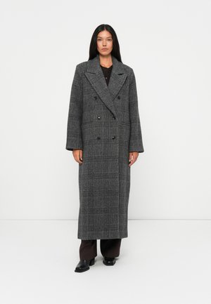 Cappotto doppiopetto grigio a quadri con grandi revers, realizzato in tessuto testurizzato, che arriva a metà polpaccio, abbinato a pantaloni scuri e stivali.