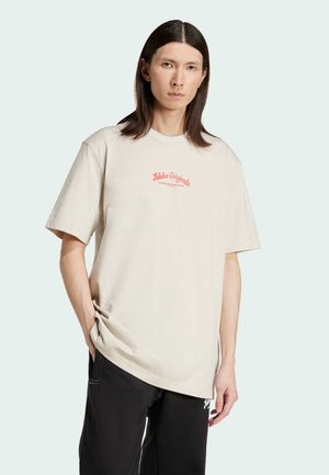 Beige oversized katoenen t-shirt met een rood logo op de borst en korte mouwen, gecombineerd met zwarte broeken. Soepele textuur, relaxte pasvorm.