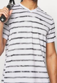 Vit träningsskjorta med svarta horisontella ränder och v-ringning. Lättviktsmaterial med en jämn yta. Liten logotyp på bröstet.