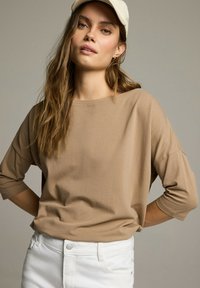 Beżowa, luźna bluzka z okrągłym dekoltem i rękawami o długości trzech czwartek, wykonana z miękkiego materiału, zestawiona z białymi, dżinsowymi spodniami o wysokim stanie.