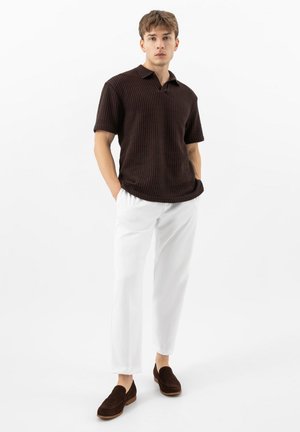 Junger Mann steht mit den Händen in den Taschen, trägt ein dunkelbraunes, kurzärmeliges Strick-Poloshirt, weiße Hosen und dunkelbraune Loafers vor weißem Hintergrund.