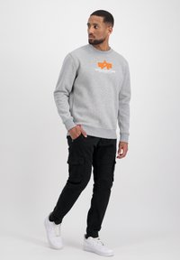 Szara bluza z pomarańczowym logo, z prążkowanymi mankietami i pasem, w połączeniu z czarnymi spodniami cargo z wieloma kieszeniami i białymi sneakersami.