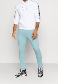 Homme portant un sweat à capuche blanc Champion avec des bandes logo sur les manches, un pantalon de jogging bleu clair, et des chaussures de sport noires, blanches et dorées, debout.