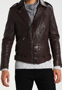 Bruine leren motorjack met zilveren ritsen, asymmetrische voorkant en een riem in de taille. Een geribbelde ronde halstrui eronder voegt textuur toe.
