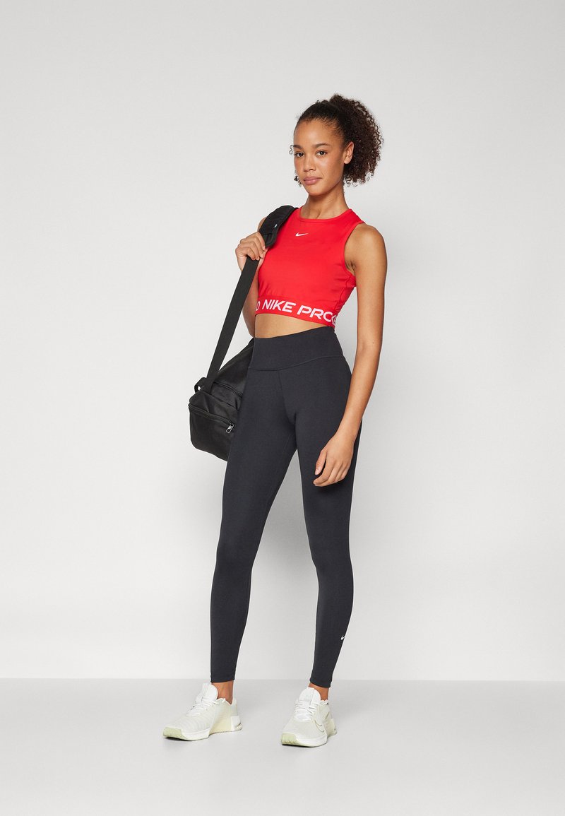 Crop top Nike Pro rouge avec logo et bande blanche, associé à des leggings noirs taille haute. Le mannequin porte un sac de sport noir et des baskets blanches.