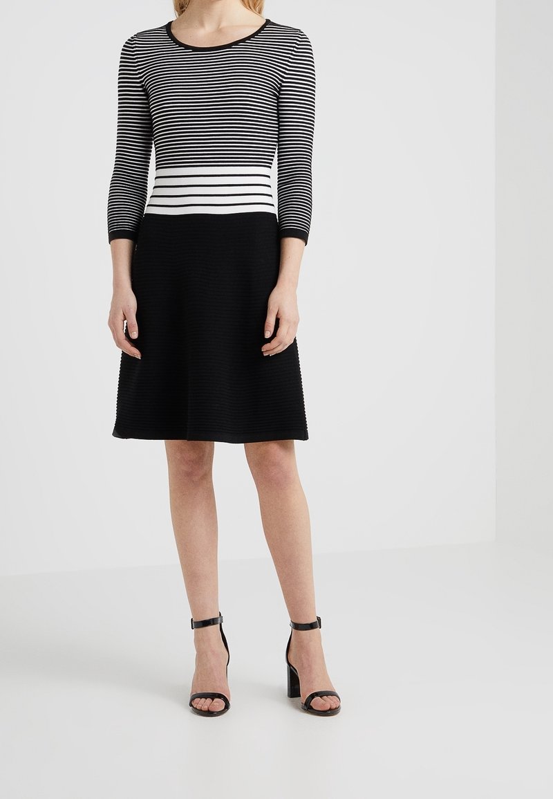 Femme portant une robe jupe noire avec un haut rayé noir et blanc et des sandales à talons hauts à bride cheville noires, se tenant sur un sol blanc.