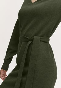 Abito a maglia verde con scollatura a V, maniche lunghe e cintura in vita. Il tessuto ha una trama morbida e una silhouette dal taglio rilassato.