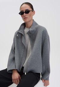 Frau mit schwarzen ovalen Sonnenbrillen, grauem Wolljacke mit Reißverschluss über einem hellgrauen Pullover und schwarzen Hosen, sitzt vor einem schlichten Hintergrund.