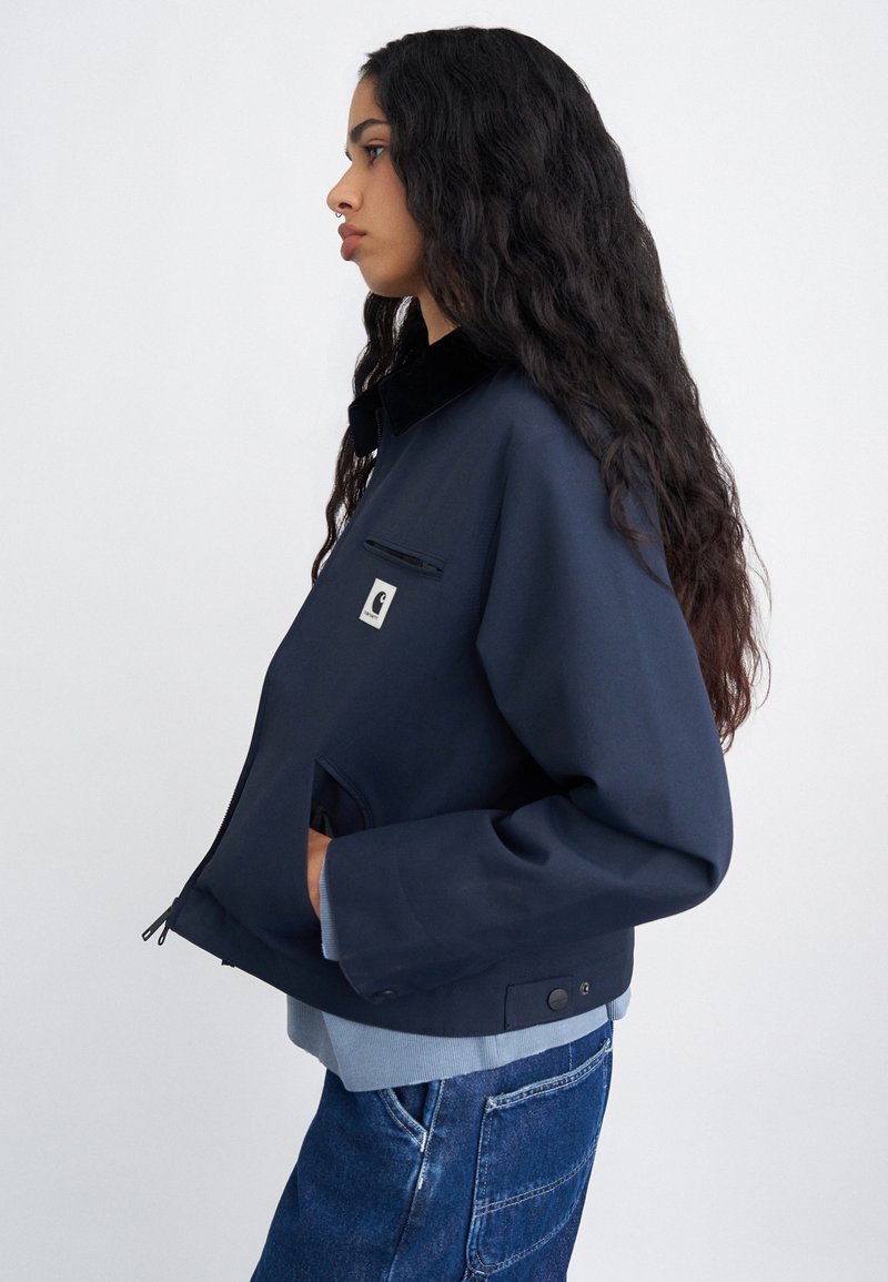 Marineblå jakke med lynlås foran, krave, brystlomme og sidelommer. Har en kontrastfarvet lyseblå kant og tekstureret stof.