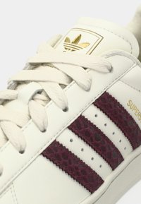 Baskets en cuir blanc avec des rayures texturées bordeaux, un logo doré sur la languette et des lacets blancs. Présente des perforations sur les côtés.