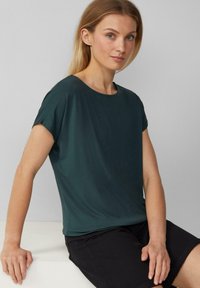 T-shirt vert foncé à manches courtes, au coupe ample, fabriqué à partir d'un tissu lisse. Il présente un col rond et un design simple. Porté avec un short noir.