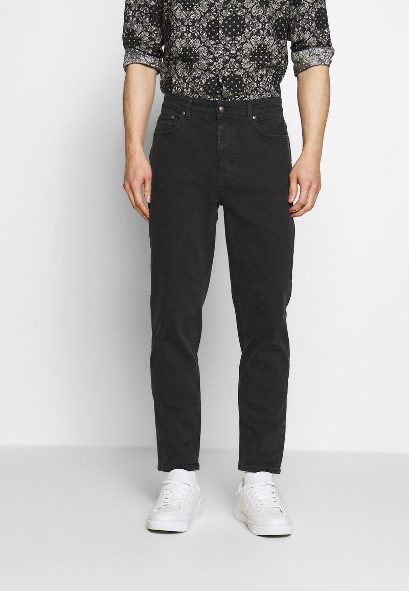 Černé džínsy s úzkým střihem, designem s pěti kapsami a jemnými detaily v švech, spárované s bílými teniskami a vzorovanou košilí.