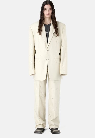 Osoba o długich ciemnych włosach, ubrana w oversize'owy beżowy garnitur z małymi czarnymi kropkami, czarny t-shirt z nadrukiem oraz czarne buty z kwadratowymi noskami.