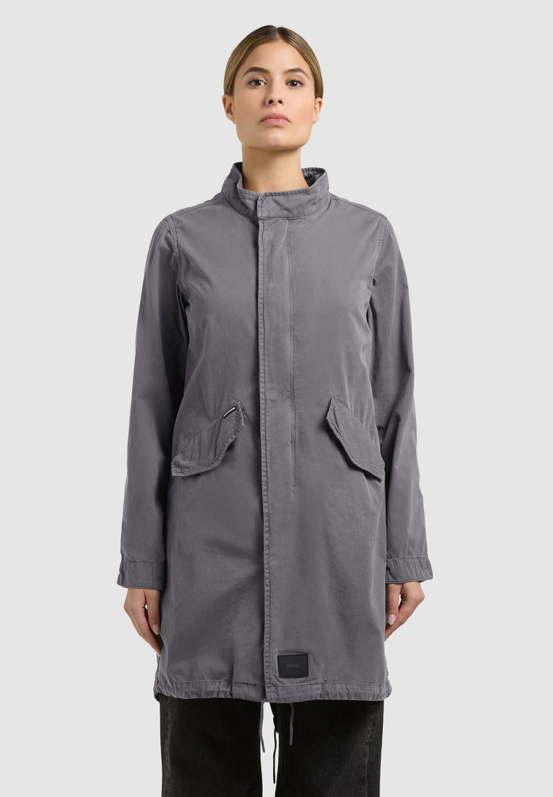 Femme portant un long manteau gris avec un col montant et des poches à rabat frontales, debout devant un fond gris clair uni.