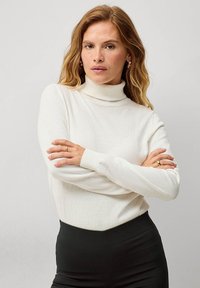 Biały sweter w stylu golf z miękkiego materiału dzianinowego, z długimi rękawami i ściągaczami, noszony z dopasowaną czarną spódnicą.