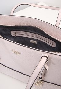 Light pink Guess handbag with double handles, embossed logo pattern, interior zip pocket, and small heart-shaped lock charm.