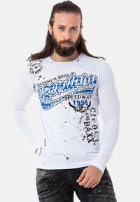 Langärmliges weißes T-Shirt mit einem blauen Grafikdruck, schwarzen Spritzern und Textdetails, kombiniert mit dunklen gemusterten Jeans.