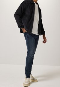 Man som bär en svart kordflodjacka, vit t-shirt, mörkblå jeans och beige sneakers, står med ena handen i fickan.