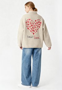 Donna con capelli castano chiaro raccolti in una coda di cavallo che indossa una giacca in pile color crema con cuori rossi e la scritta "SWEET heart", abbinata a jeans larghi blu e scarpe bianche.