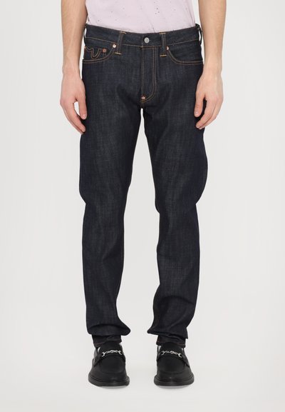 Jeans din denim de culoare bleumarin închis, cu o croială dreaptă, cusături portocalii în contrast și detalii ale buzunarelor din față. Purtați cu pantofi negri.