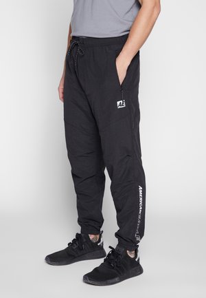 Mężczyzna noszący czarne spodnie dresowe American Eagle z sznurkiem w pasie i zamykanymi na zamek kieszeniami, w połączeniu z czarnymi sneakersami Adidas.