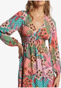 Eine Frau trägt ein farbenfrohes, geblümtes Patchworkkleid mit langen Ärmeln und einem tiefen V-Ausschnitt, ergänzt durch geschichtete Goldketten.