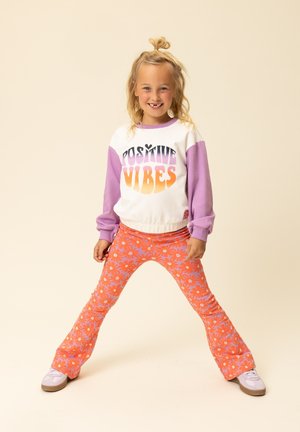 Sweatshirt met een wit lichaam en paarse mouwen, met de tekst "Positive Vibes". Wijde oranje broek met wit bloemenpatroon. Witte sneakers.