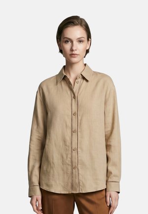 Giovane donna che indossa una camicia beige a maniche lunghe con bottoni e pantaloni marroni, in piedi davanti a uno sfondo chiaro e semplice.